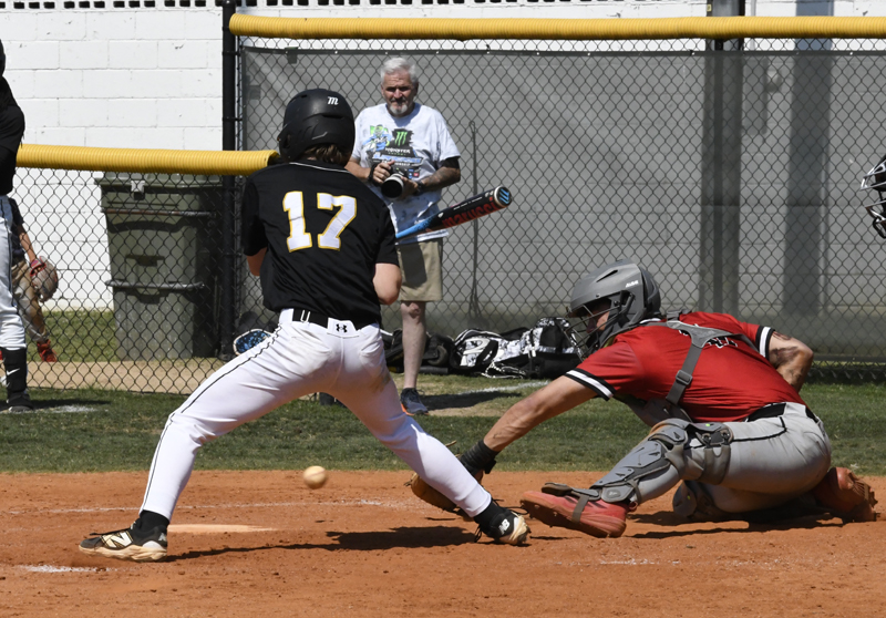 A baseball player in black dodges a pitch while the catcher in red attempts to catch the ball; a spectator with a camera watches in the background.