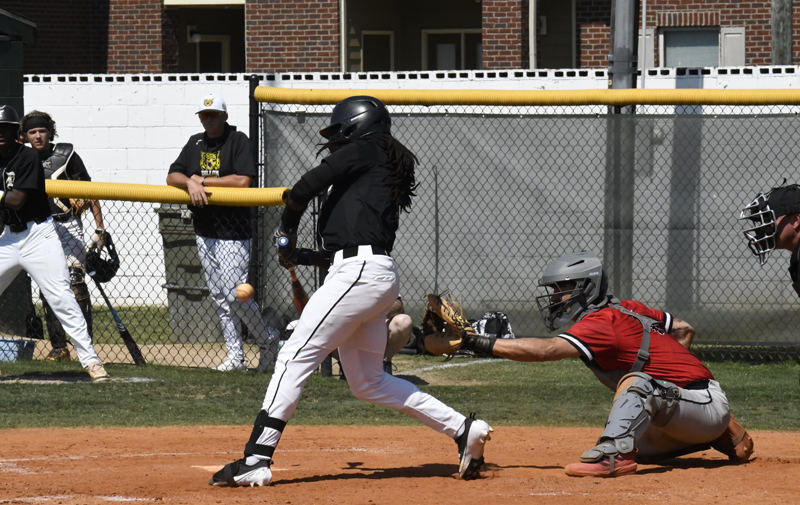 Baseball player in black uniform swinging bat at pitch, catcher in red gear ready to catch ball, other players and a fence in the background.