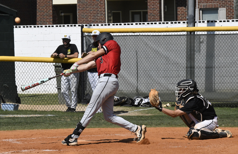 A baseball player in a red jersey swings at a pitch while a catcher and spectators watch from behind.