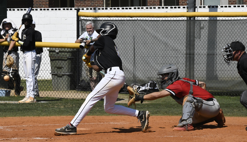 A baseball player in black swings at a pitch as the catcher and umpire watch closely; teammates and spectators are in the background.