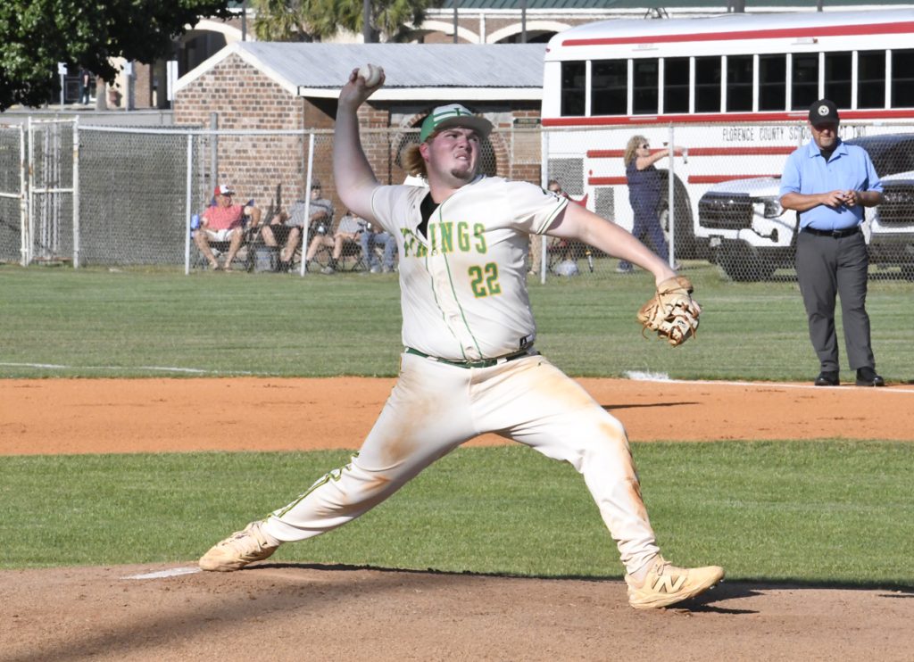 Baseball pitcher in uniform mid-throw on a sunny field, with spectators and an umpire in the background.