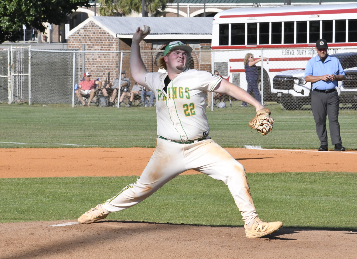 Baseball pitcher in uniform mid-throw on a sunny field, with spectators and an umpire in the background.
