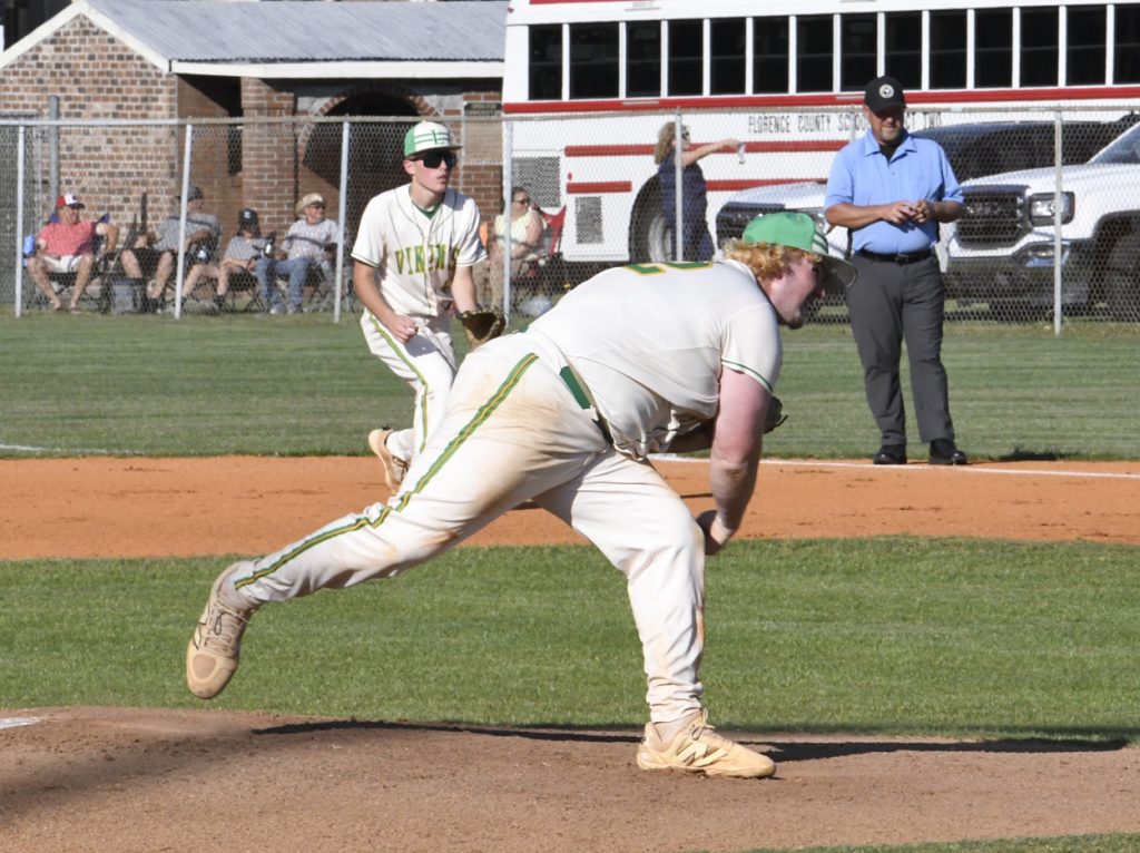 Baseball pitcher in mid-throw with teammate and umpire in background on a sunny field, spectators watching behind a fence.