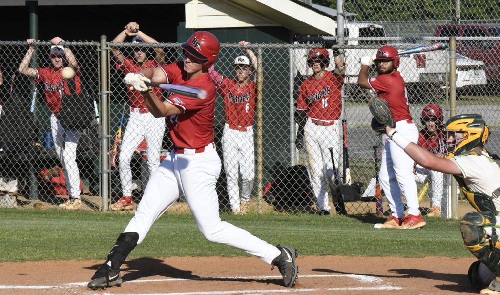 Baseball player in red uniform swinging bat at a pitch, with teammates and a catcher visible, during a game on a sunny day.