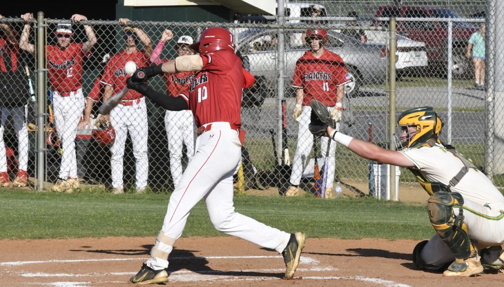 Baseball player in red swings at a pitch, while the catcher in white and green gear prepares to catch the ball; teammates watch from behind the fence.