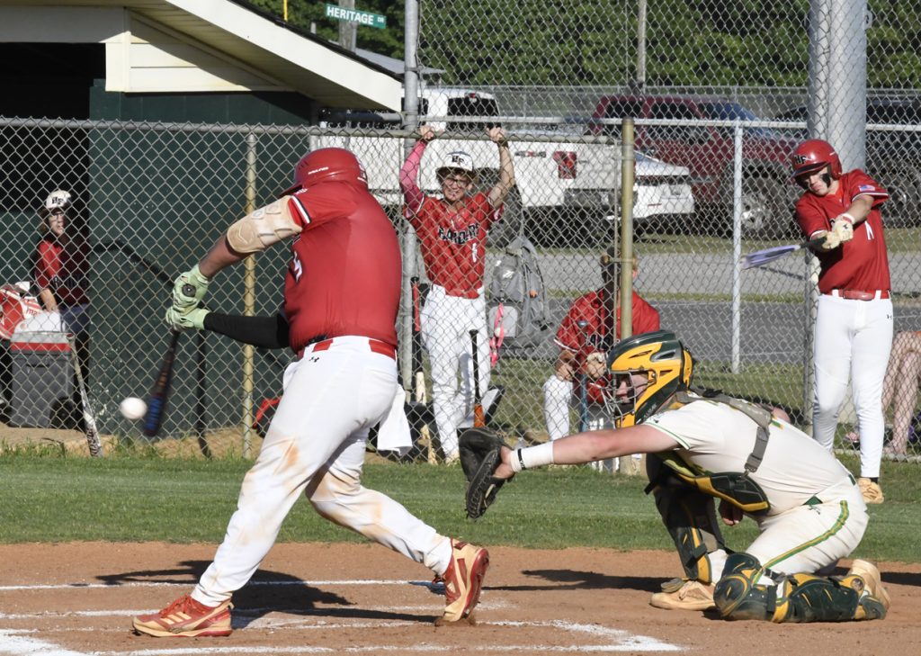 Baseball player in red uniform swinging bat at a pitched ball, catcher in green gear, teammates and spectators in the background behind a chain-link fence.
