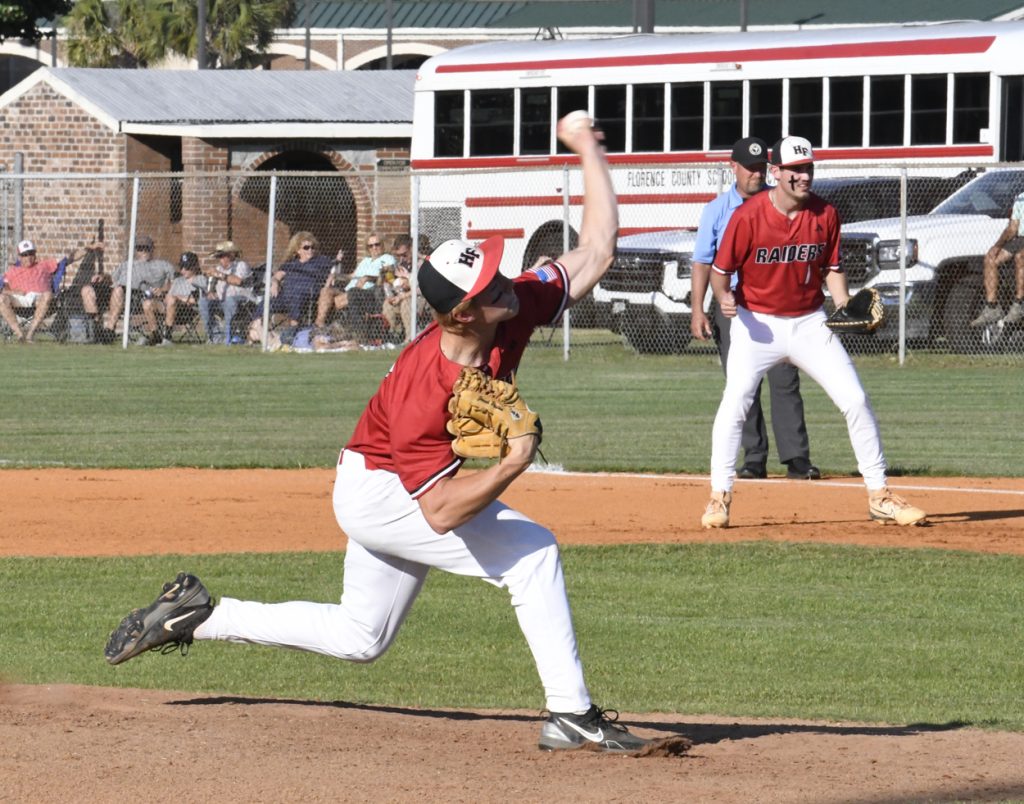 A baseball pitcher in mid-throw on a field, with teammates, umpire, spectators, and a school bus in the background.