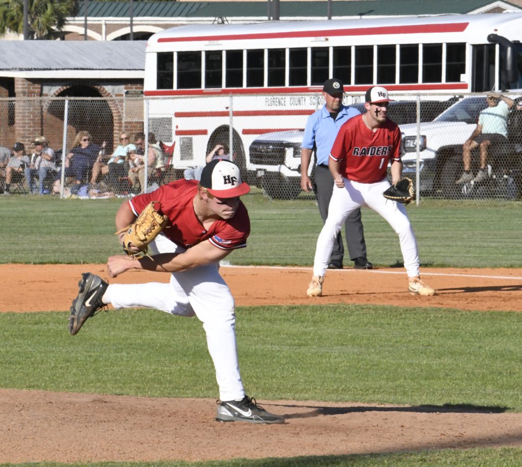 Baseball pitcher in red and white uniform throwing a pitch, with teammate, umpire, and school bus in the background on a sunny day.