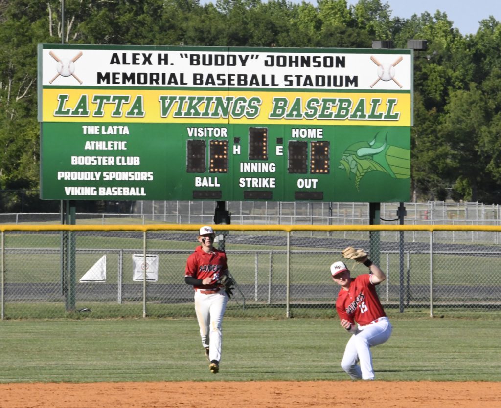 Two baseball players in red jerseys on a field with a scoreboard in the background reading “Latta Vikings Baseball.”