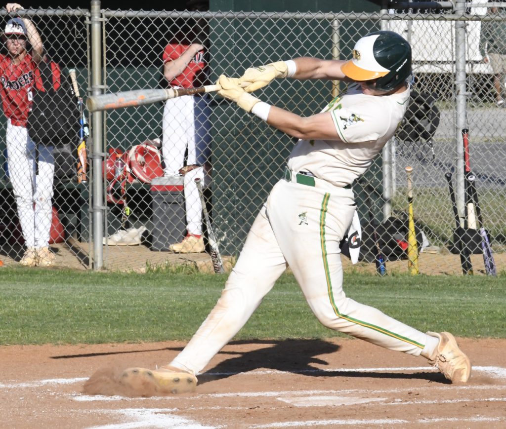 Baseball player in white uniform swinging bat at pitched ball during a game, with teammates and fence visible in the background.