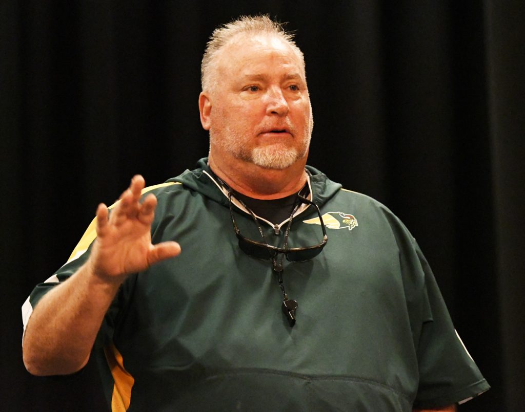 A man in a green sports shirt with sunglasses and a whistle around his neck gestures while speaking indoors against a dark background.