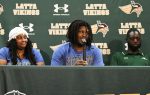 Three people sit at a press conference table with "Latta Vikings" and Under Armour logos on a green backdrop. The center person speaks into a microphone.