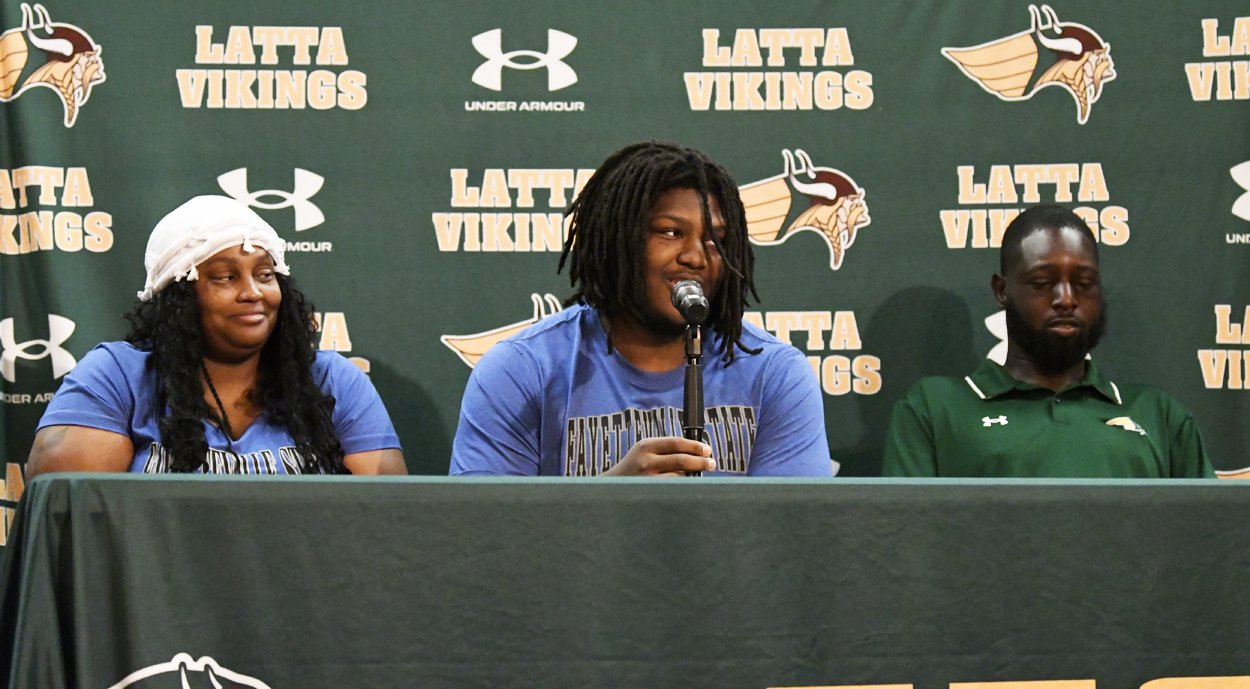 Three people sit at a press conference table with "Latta Vikings" and Under Armour logos on a green backdrop. The center person speaks into a microphone.
