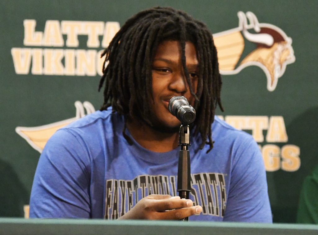 A person with dreadlocks speaks into a microphone, wearing a blue "Fayetteville State" shirt, sitting in front of a "Latta Vikings" backdrop.