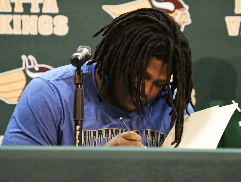 A person with dreadlocks sits at a table, signing a document with a pen, microphone nearby, in front of a green backdrop with "Vikings" text.