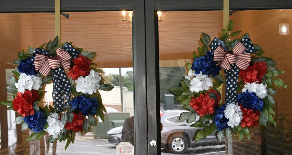 Two glass doors decorated with patriotic wreaths featuring red, white, and blue flowers, greenery, and small American flags. A car and street are visible outside.