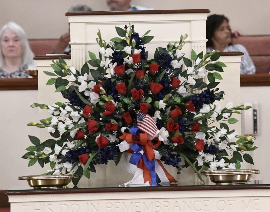 Large patriotic floral arrangement with red, white, and blue flowers, accented by small American flags and ribbons, displayed indoors with people in the background.