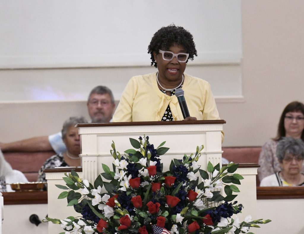 A woman speaks at a podium decorated with red, white, and blue flowers in a church; people are seated in the background.
