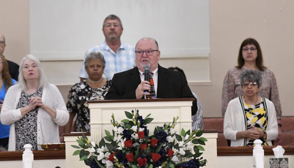 A man in a suit speaks at a church pulpit with a microphone, while several people stand behind him. Floral arrangement is on the pulpit.