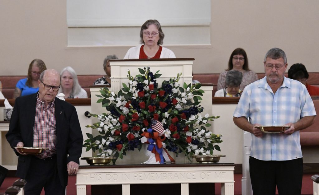 A woman stands behind a podium with a large floral arrangement, surrounded by seated people in a church setting.