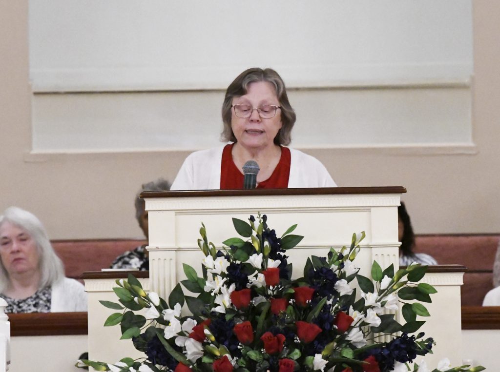 A woman stands at a podium with a microphone, behind a large floral arrangement, speaking in a church or formal indoor setting.
