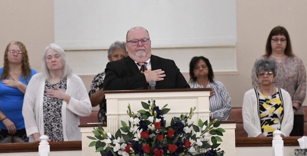 A man in a suit stands at a podium with hand on chest, flowers in front, and three women in the background inside a church.