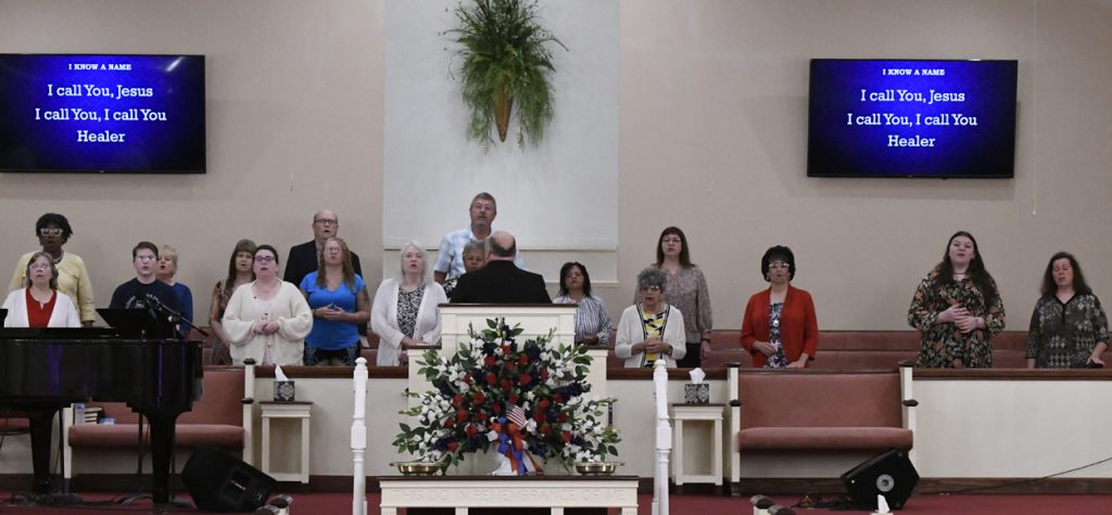 A group of people standing and sitting at the front of a church sanctuary, behind a pulpit and floral arrangement.