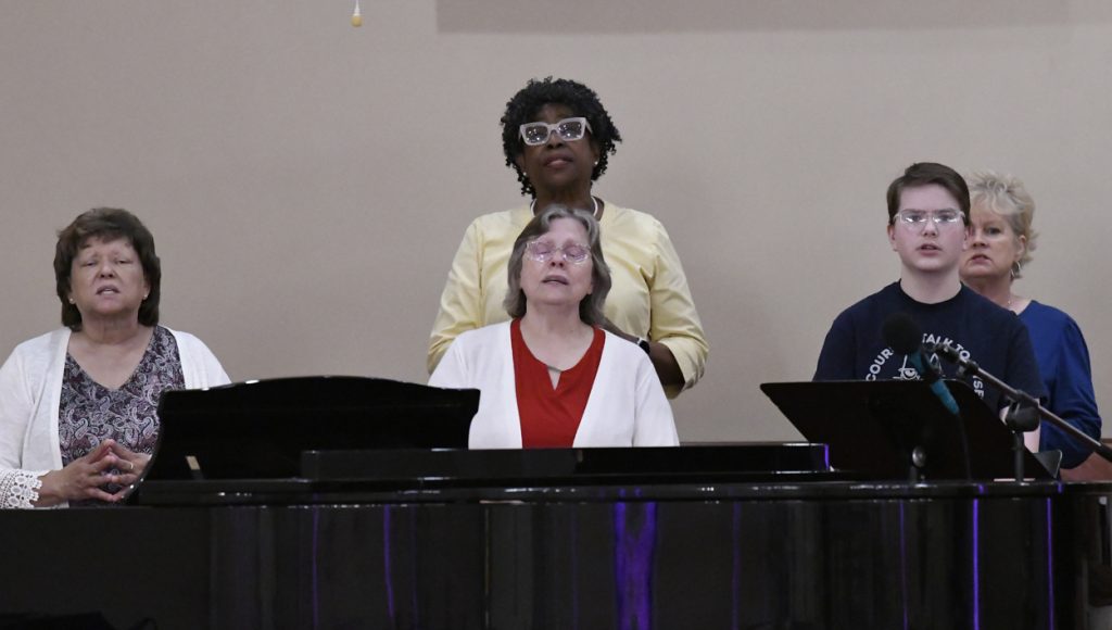 Two women are standing behind a piano, singing or performing in what appears to be a church or music recital setting.