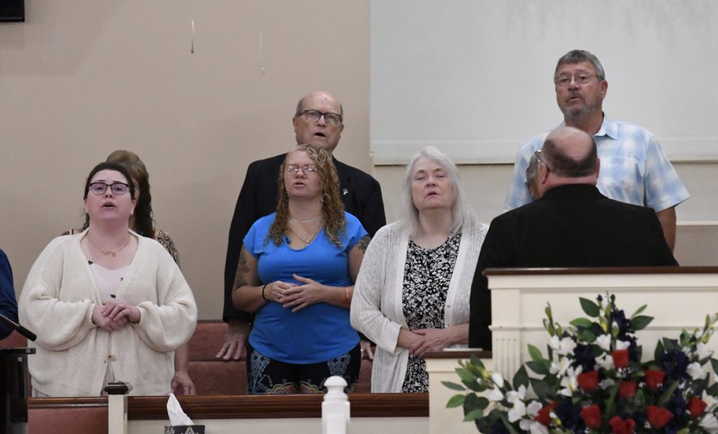 A group of people standing and singing in a church, some with eyes closed, in front of a pulpit decorated with flowers.