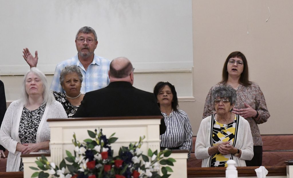 A group of people singing in a church choir, standing behind a pulpit decorated with red, white, and blue flowers.