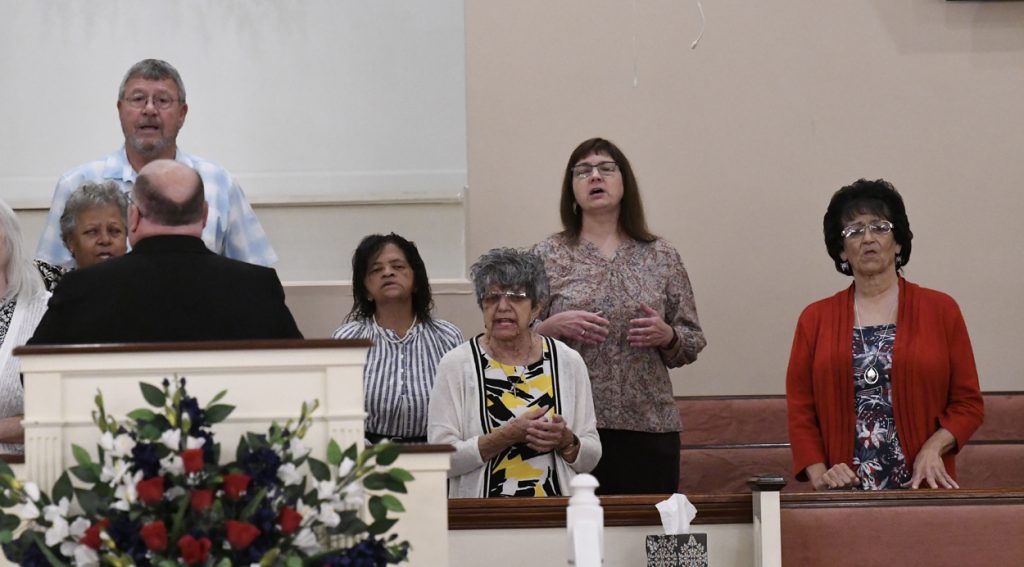 Four women standing and singing in a church, with flower arrangements and white pillars visible in the foreground.