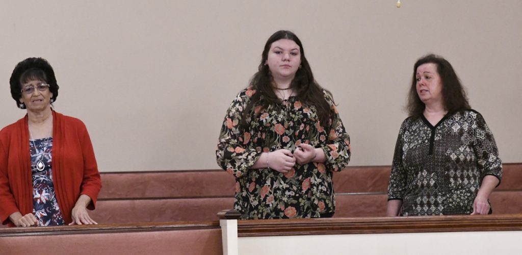 A woman with long brown hair wearing a floral dress stands indoors in front of wooden pews and a beige wall.
