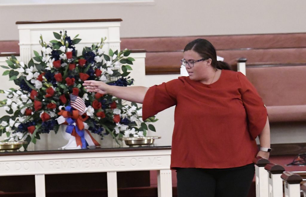 A woman in a red top gestures toward a floral arrangement with American flags on a table inside a building.