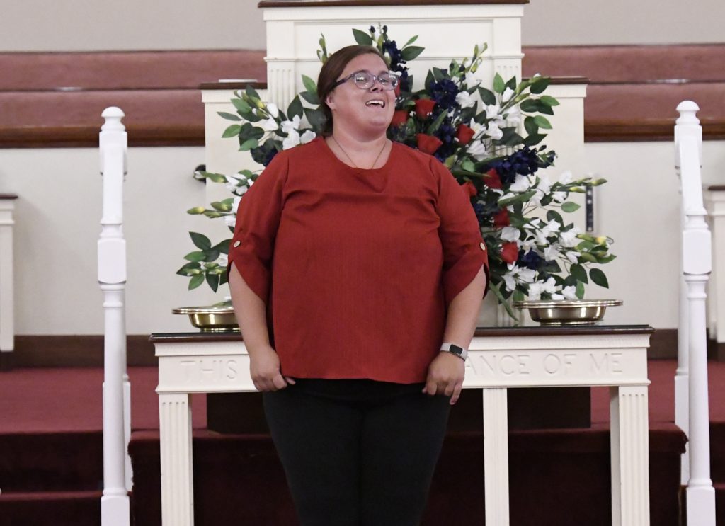 A woman in a red top stands in front of a decorated table with flowers, smiling and looking upwards indoors.