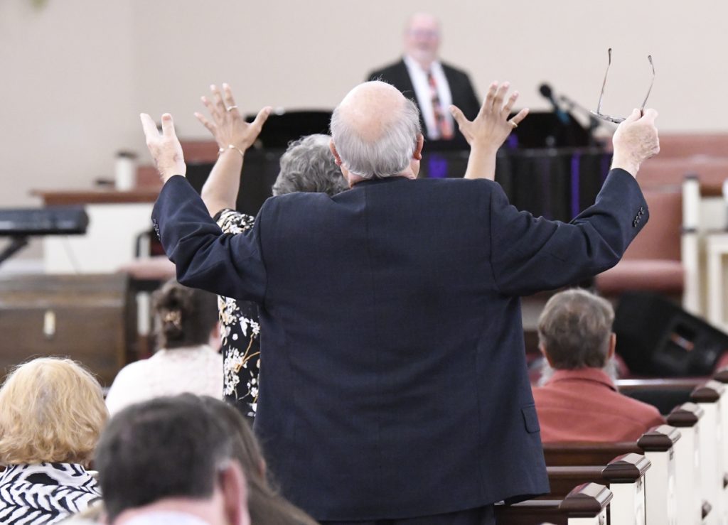 People in a church pew raising their hands in worship as a preacher speaks at the front of the room.