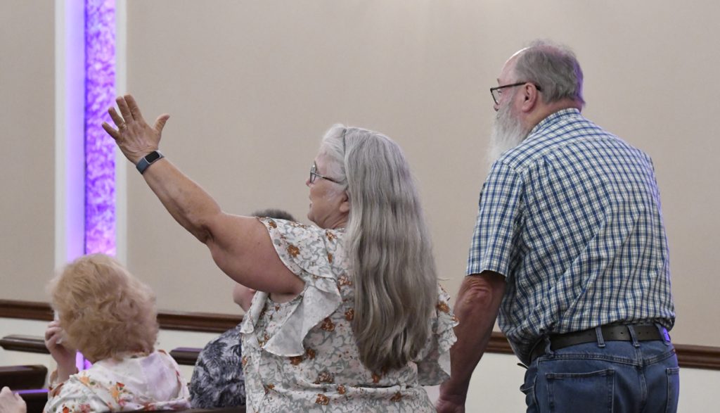 older woman with long gray hair raises her arm while standing next to an older man with a beard in a casual indoor setting.