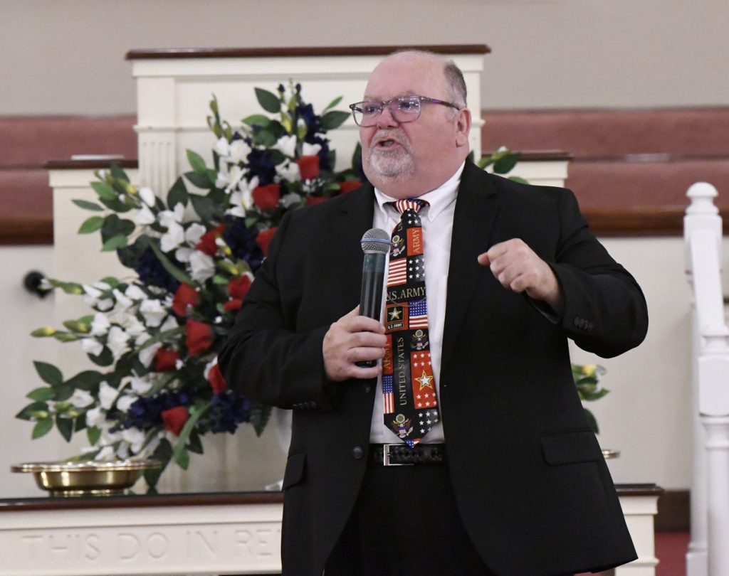 A man in a suit and colorful tie speaks into a microphone in a church, with floral arrangements in the background.