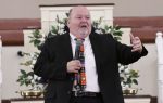 A man in a black suit speaks into a microphone, gesturing with one hand, standing in front of floral arrangements indoors.