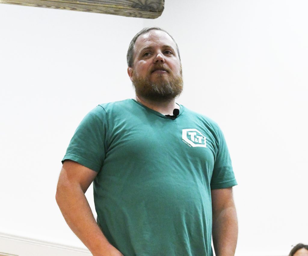 A bearded man in a green t-shirt stands indoors, looking slightly upwards, with a white background and wooden beam visible above.