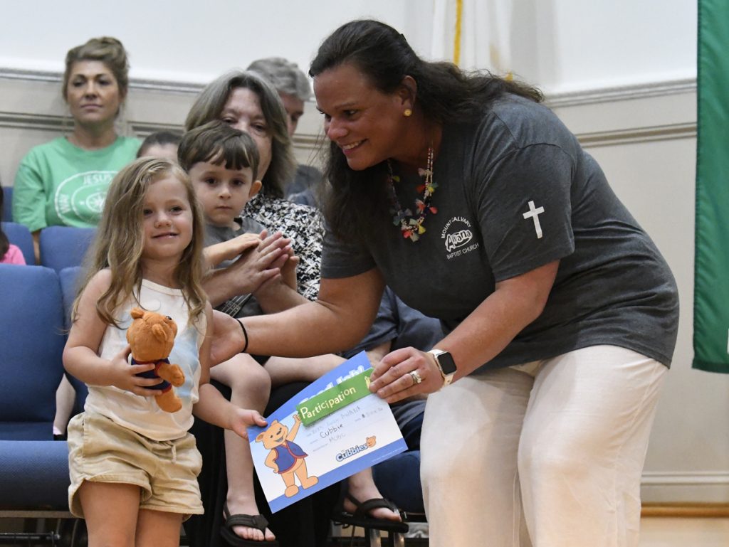 A woman hands a certificate to a young girl holding a teddy bear, while people watch and smile in the background at an indoor event.