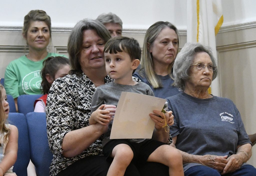 A woman holds a young boy with short hair and a paper, sitting among a group of adults and seniors at an indoor event.
