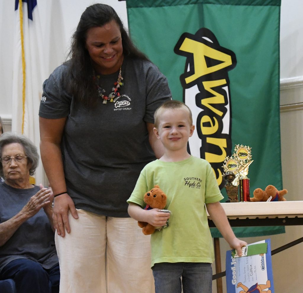 A woman and a child stand together at an awards event; the child holds a certificate and toy, with a green "Awards" banner in the background.
