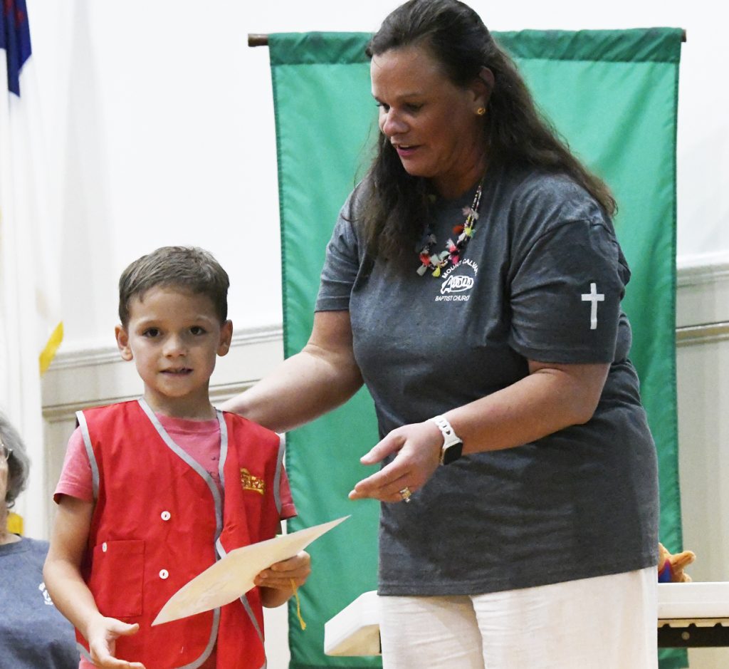 A woman hands a certificate to a young boy wearing a red vest, both smiling during an indoor award ceremony.