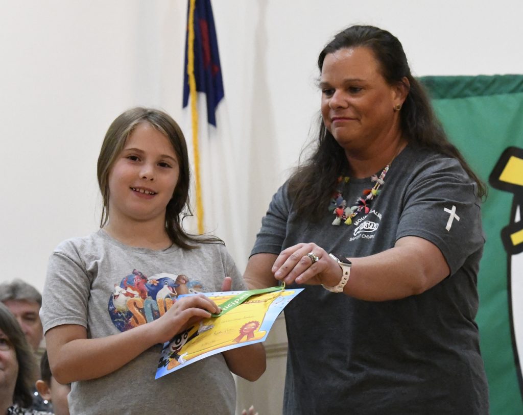 A woman and a young girl smiling while holding a colorful certificate together indoors, with a flag and green cloth in the background.