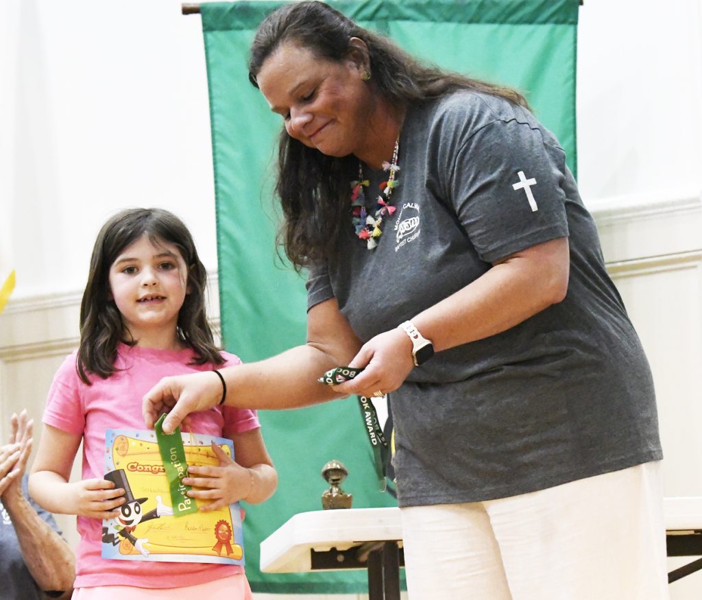 A woman hands an object to a young girl holding a book, both standing indoors with a green banner in the background.