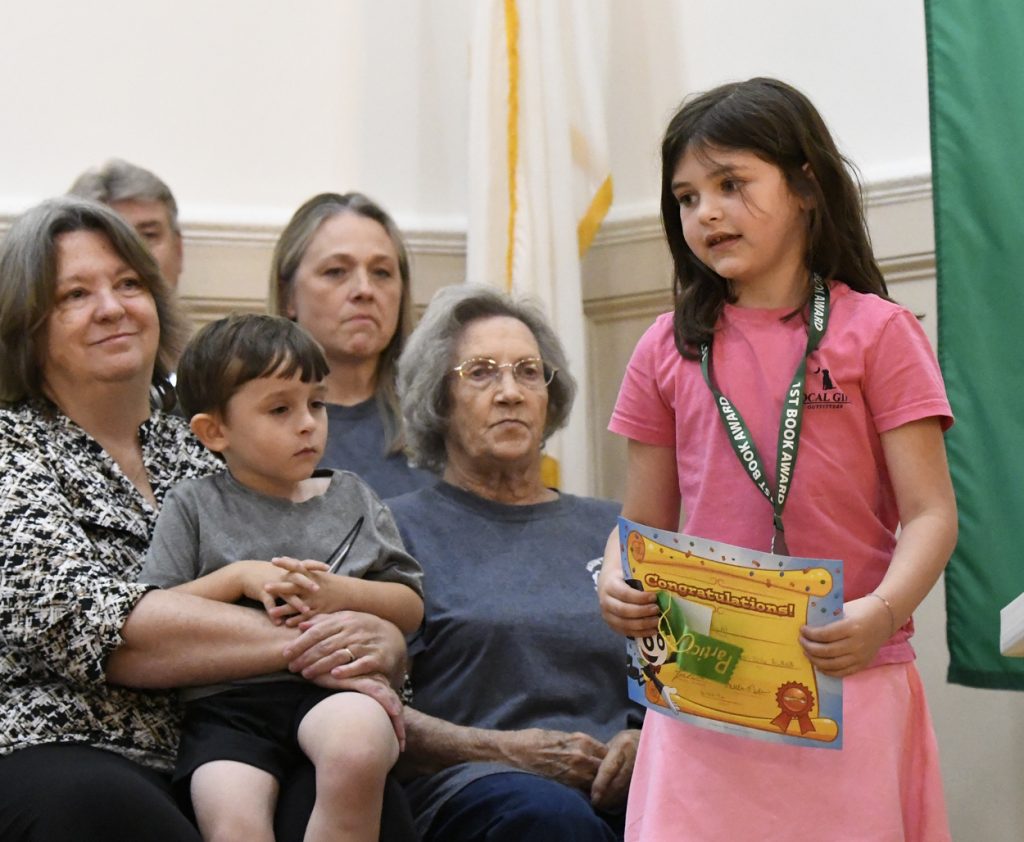 A young girl stands holding a certificate while a group of adults and a child watch her during an indoor event.