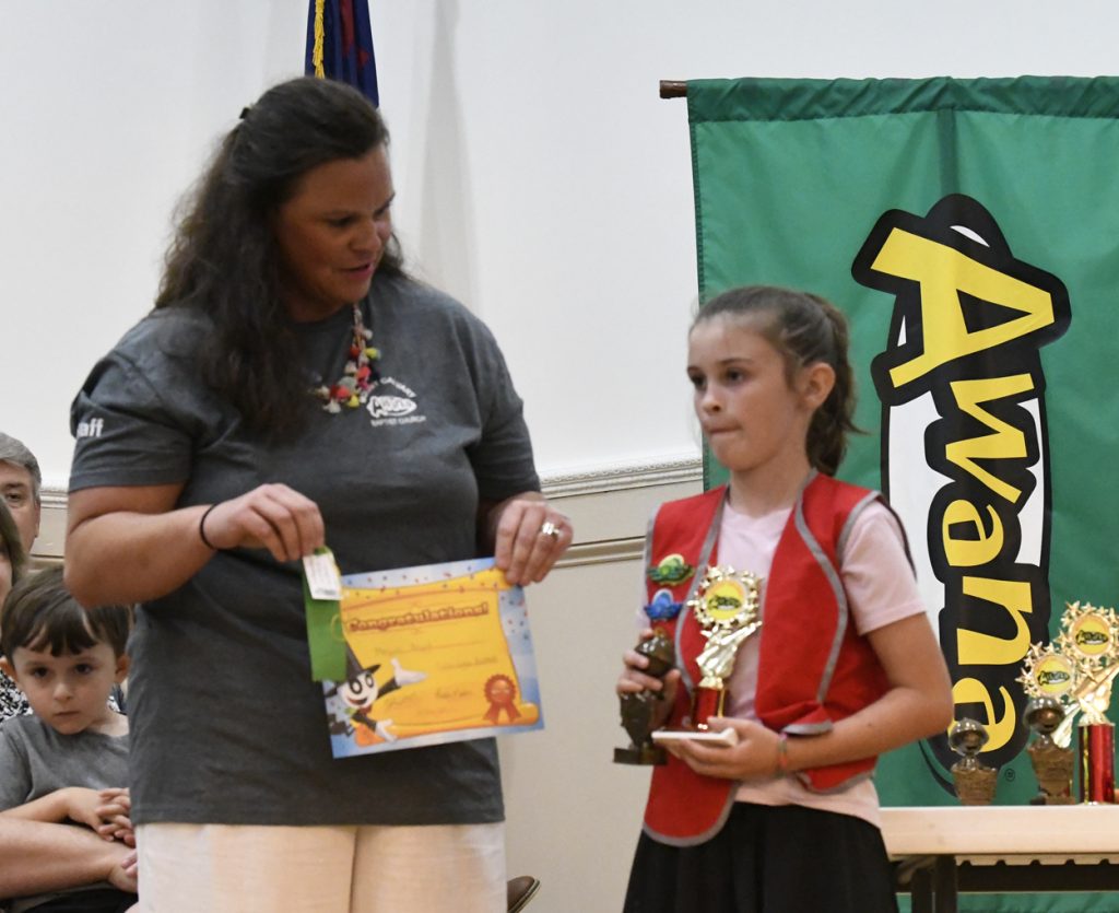 A woman hands a certificate and ribbon to a young girl holding a trophy at an awards ceremony.