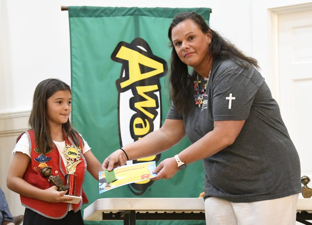 A woman hands a certificate to a girl holding a trophy, standing in front of a green "Awards" banner.
