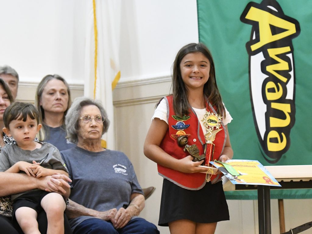Smiling girl in a red vest holding a trophy stands near a table, with people seated behind her and a green banner in the background.