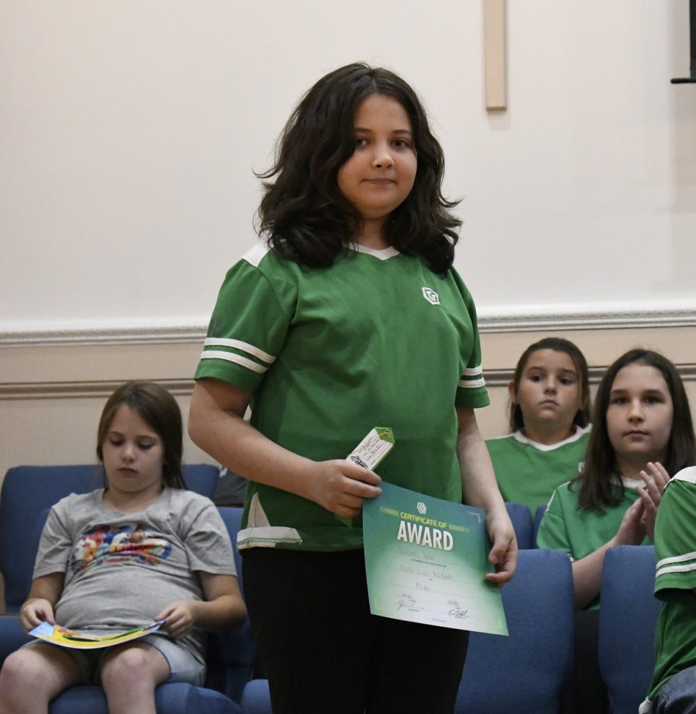 A girl in a green shirt stands holding a certificate, while other children sit on blue chairs in the background.